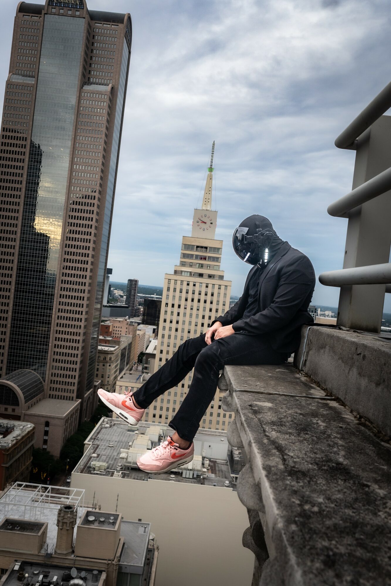 Person sitting on rooftop ledge overlooking city skyline with tall buildings and cloudy sky.