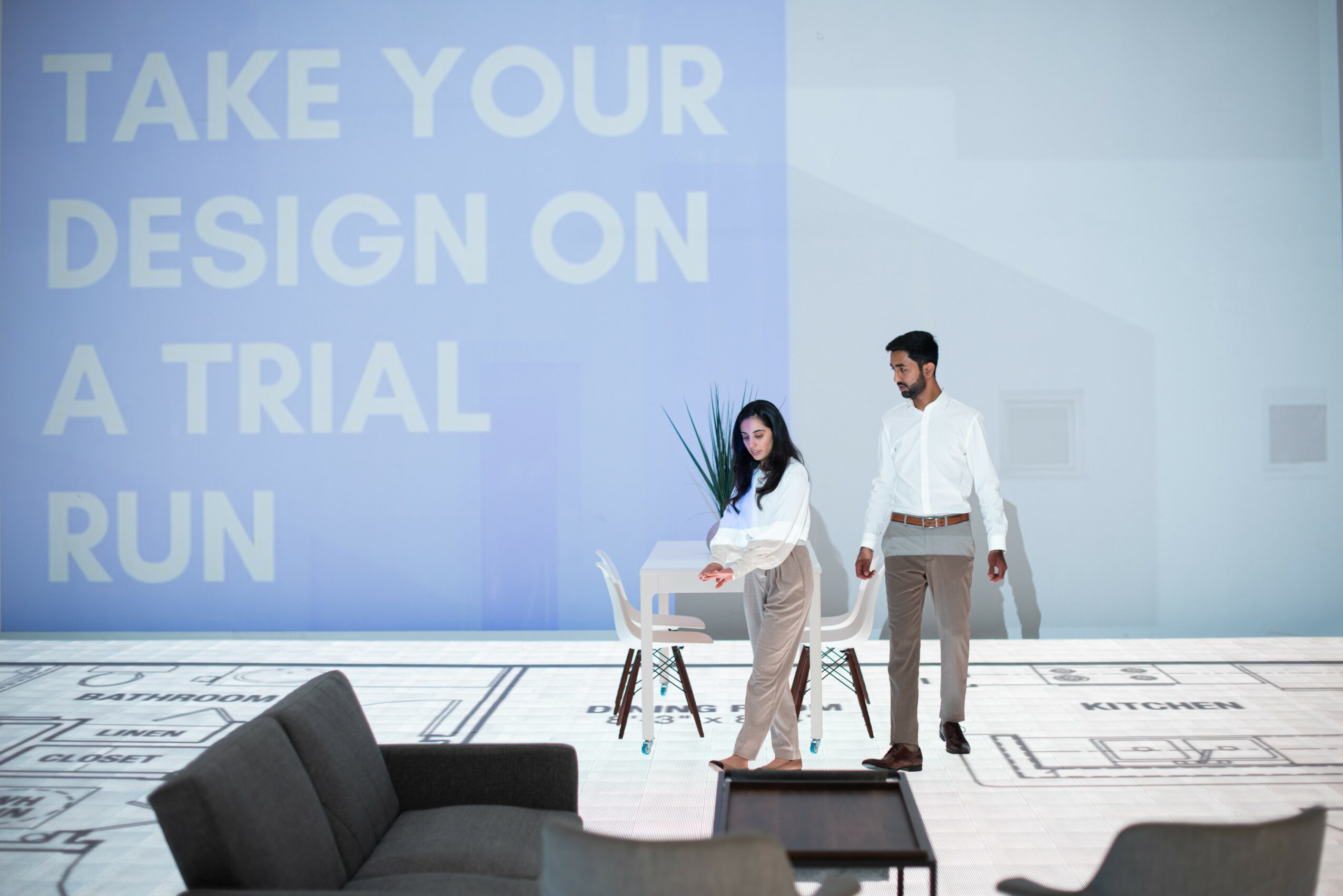Two people walking in a modern office with a large screen displaying text, and a black sofa in the foreground.