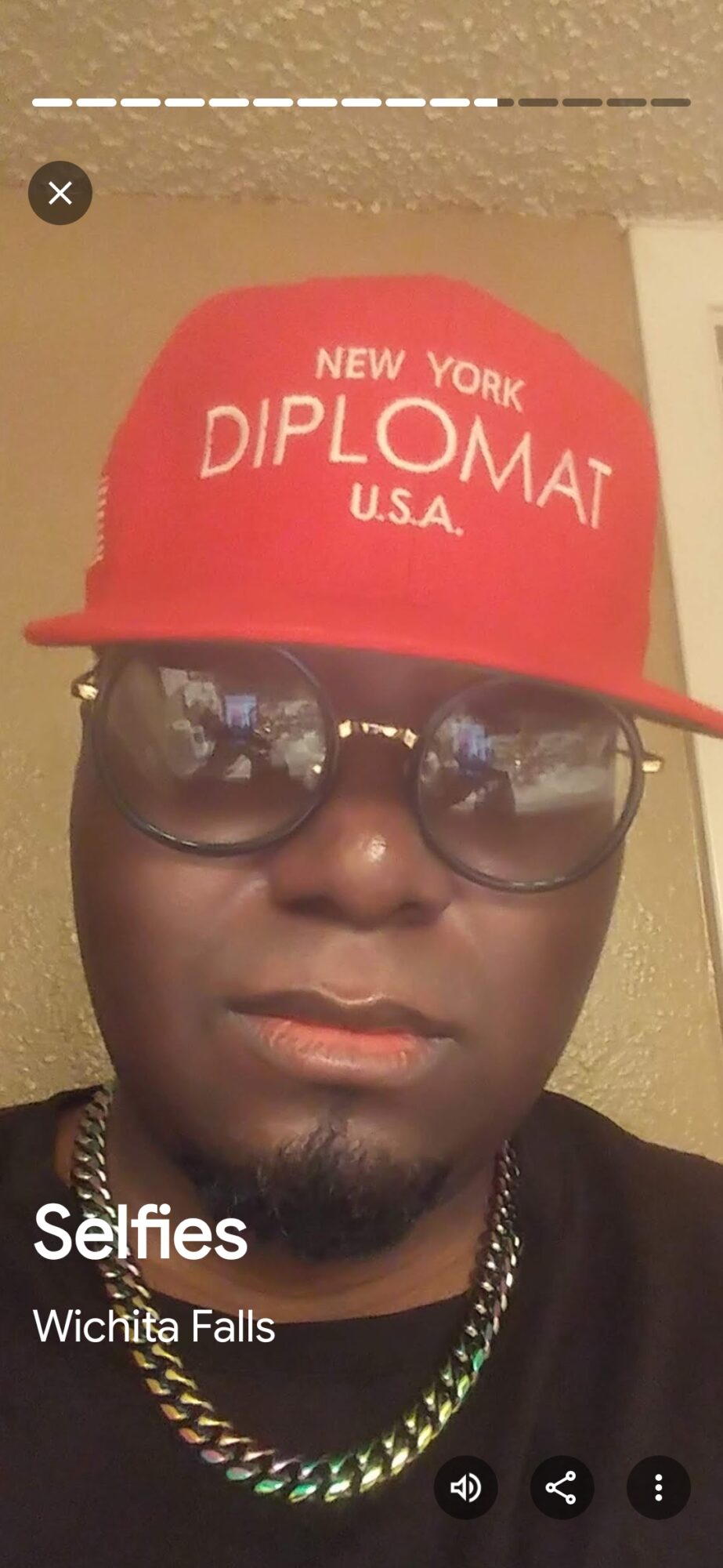 Person wearing glasses and a red cap with white text, indoors, with a chain necklace, looking at the camera.