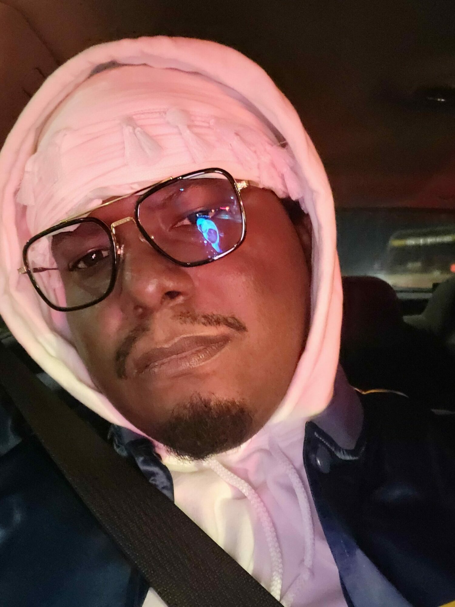 Person wearing glasses and a pink hoodie, lying down with eyes closed, in a dimly lit environment.