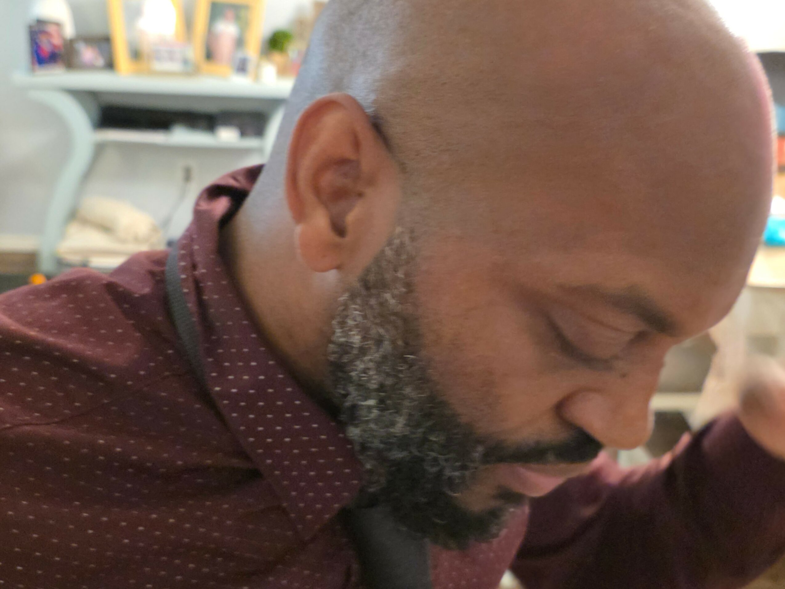 Close-up of a man with a shaved head and beard, looking downward, wearing a maroon shirt with small white dots.