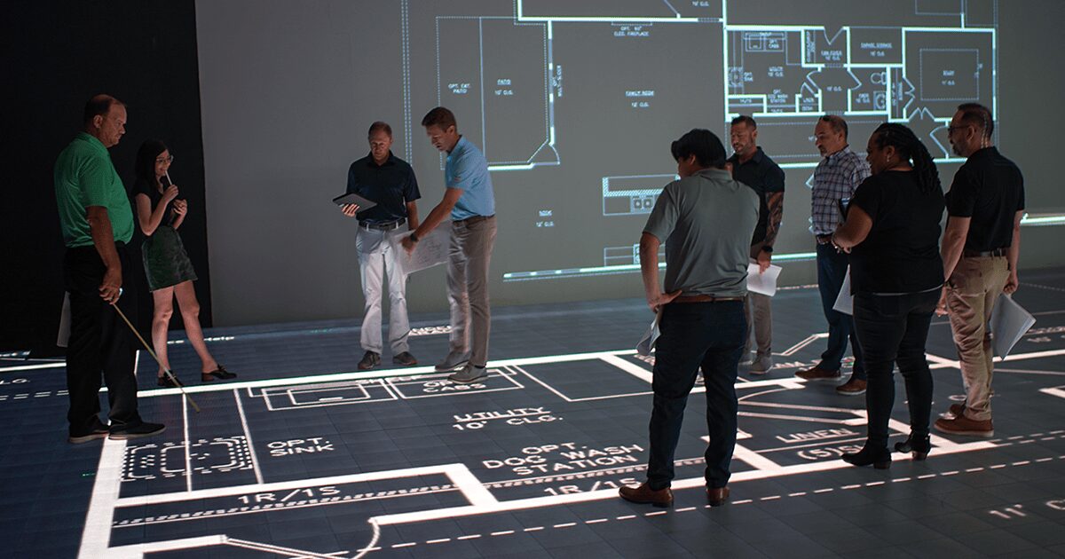 Group of people standing around a large floor plan and a projected blueprint on the wall, discussing the design.