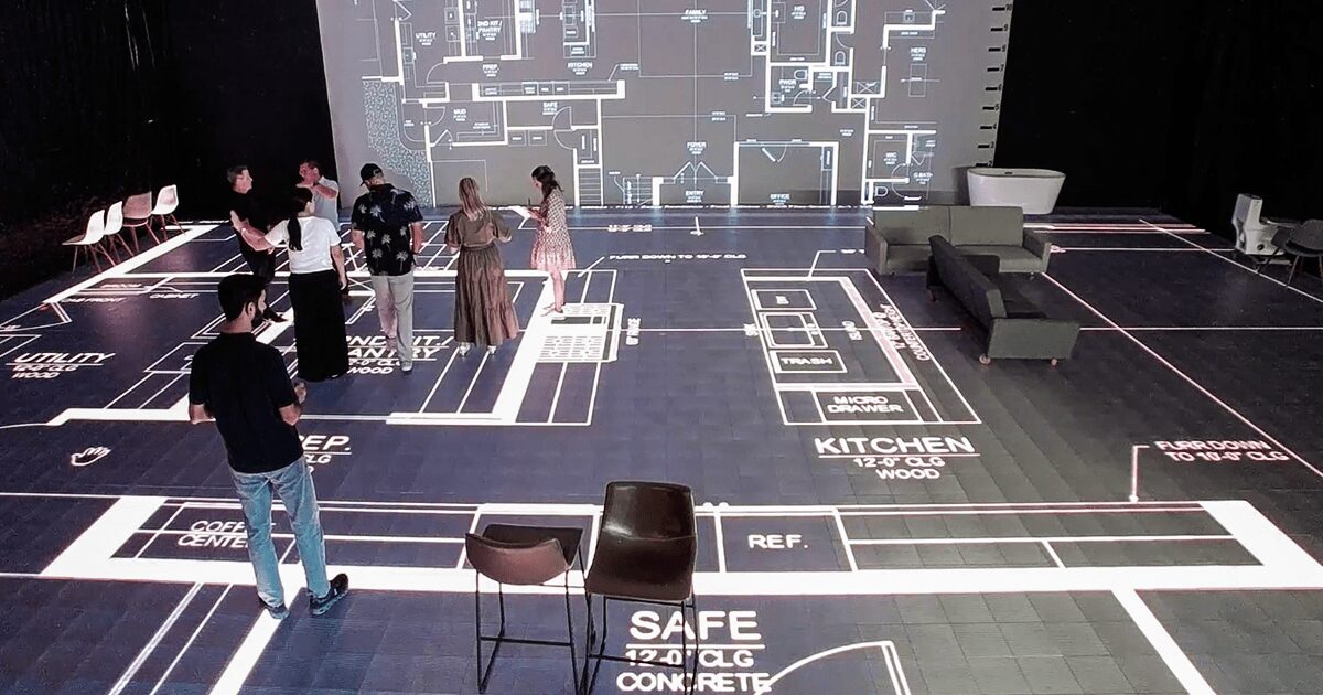 People in a room with floor plans and diagrams projected on the floor and wall, some standing and observing, with chairs and furniture.