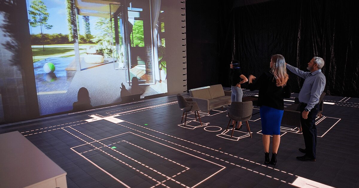 Three people stand on a stage with a large screen displaying a park scene, and floor markings are visible.