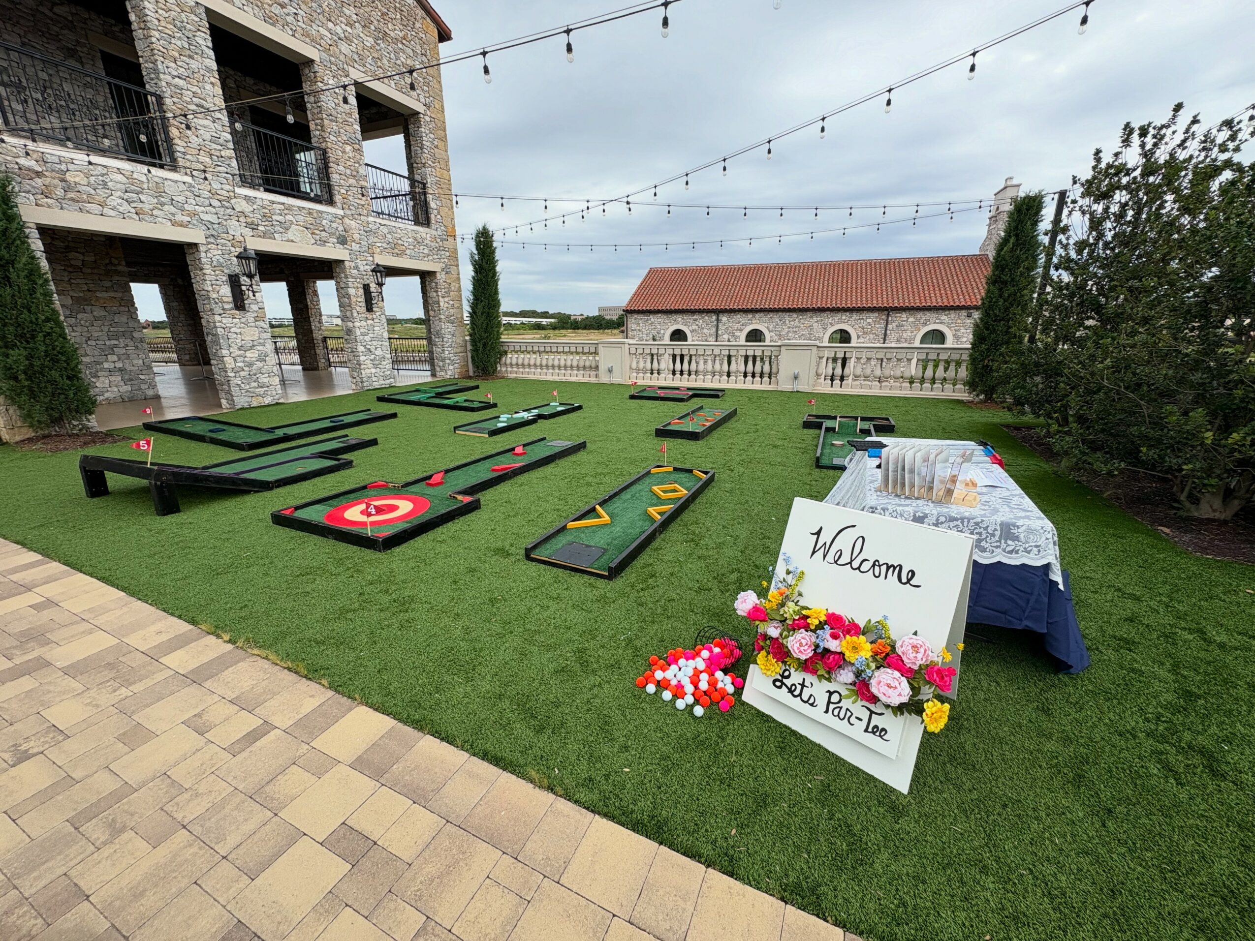 Miniature golf course with various holes on a grassy area, a welcome sign, and a table with flowers and a sign, outdoors.