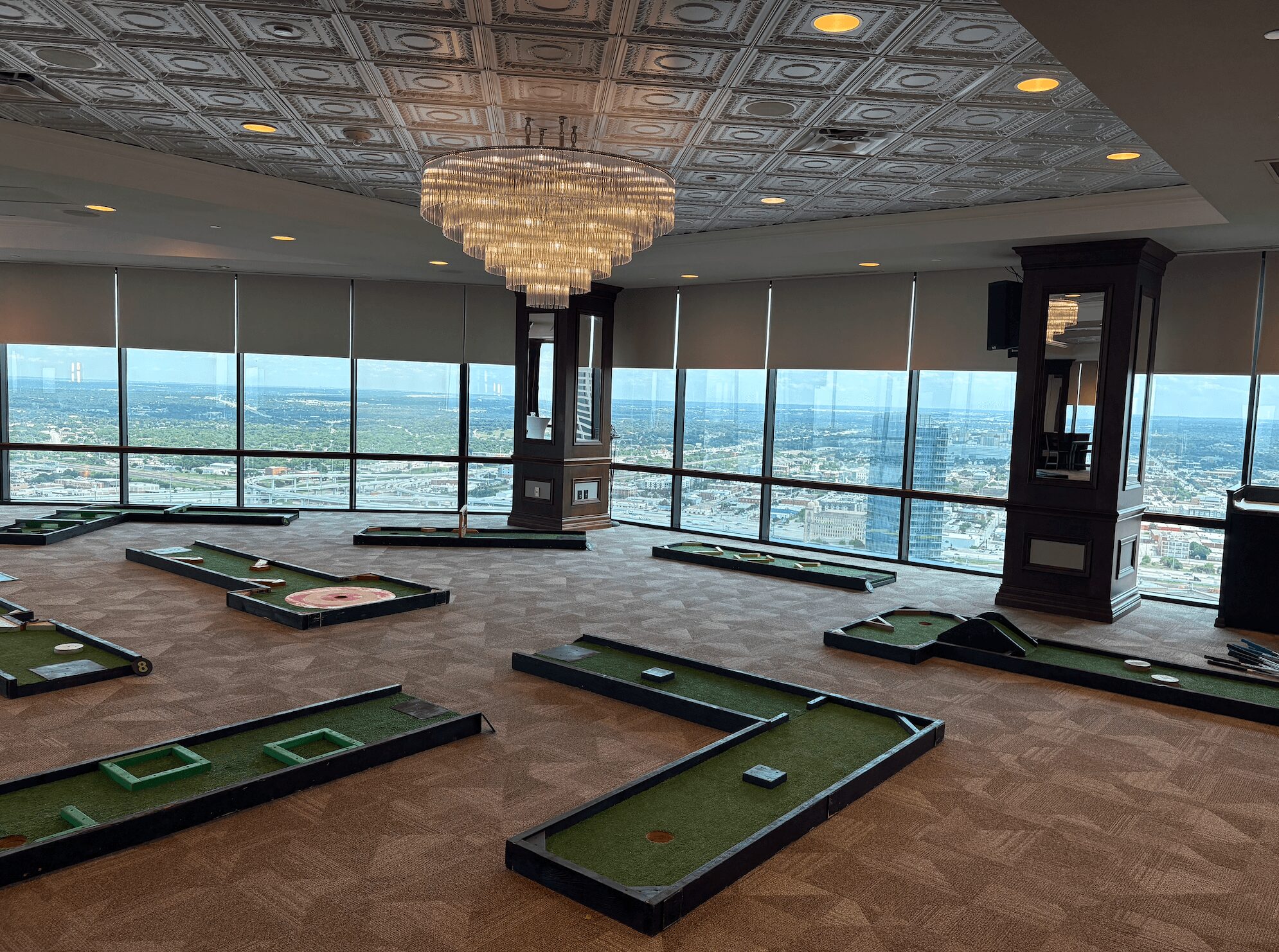 Indoor mini golf course with multiple holes, large windows, and a chandelier, overlooking a cityscape. Word count: 20.