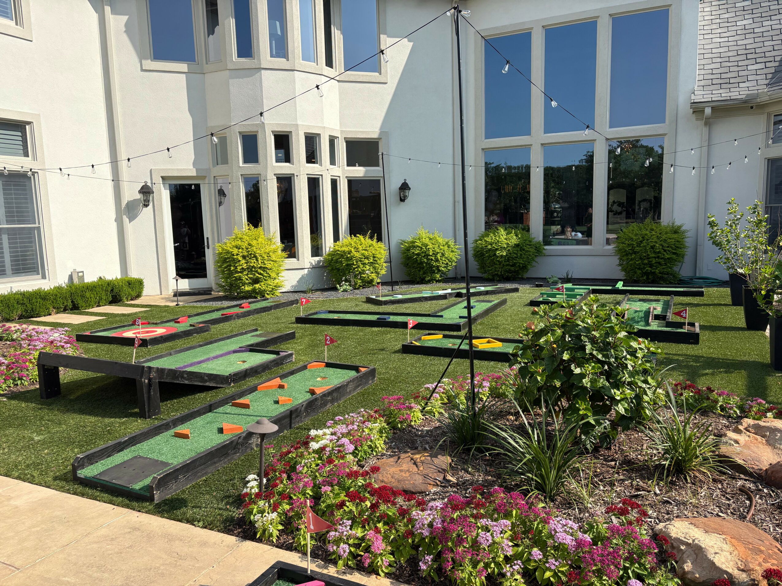 Miniature golf course with multiple holes, lush green grass, and colorful flowers in front of a modern building.