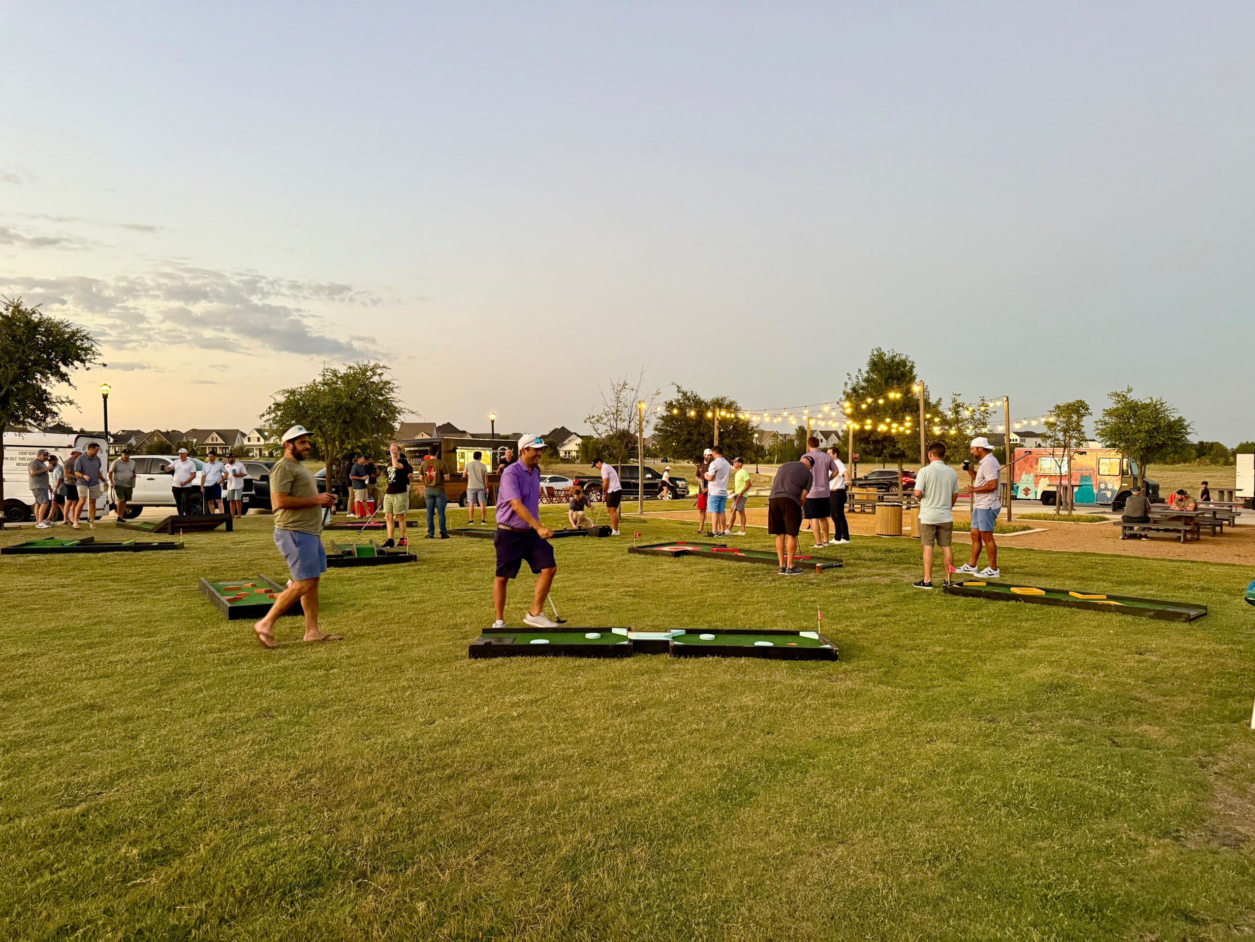 People gather outdoors on a grassy field during daytime, some walking and others standing, with trees and vehicles in the background.
