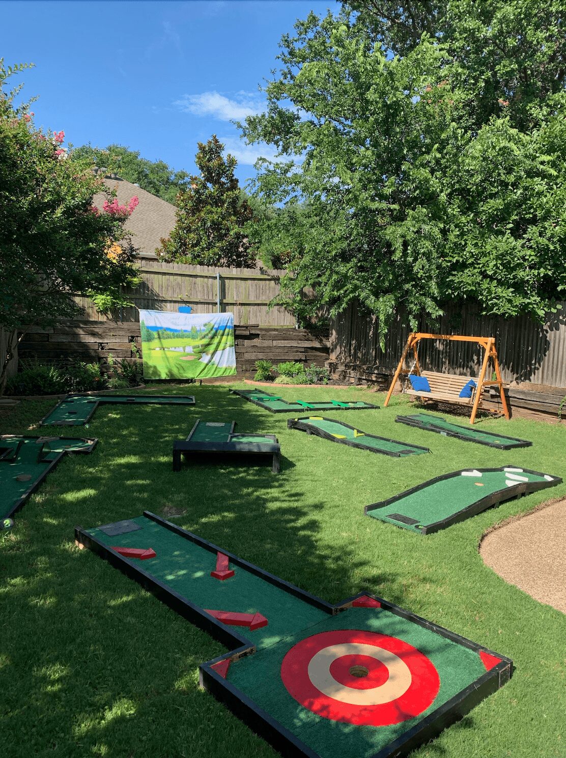 Miniature golf course with multiple holes, a target, and a swing set in a backyard with trees and a wooden fence.