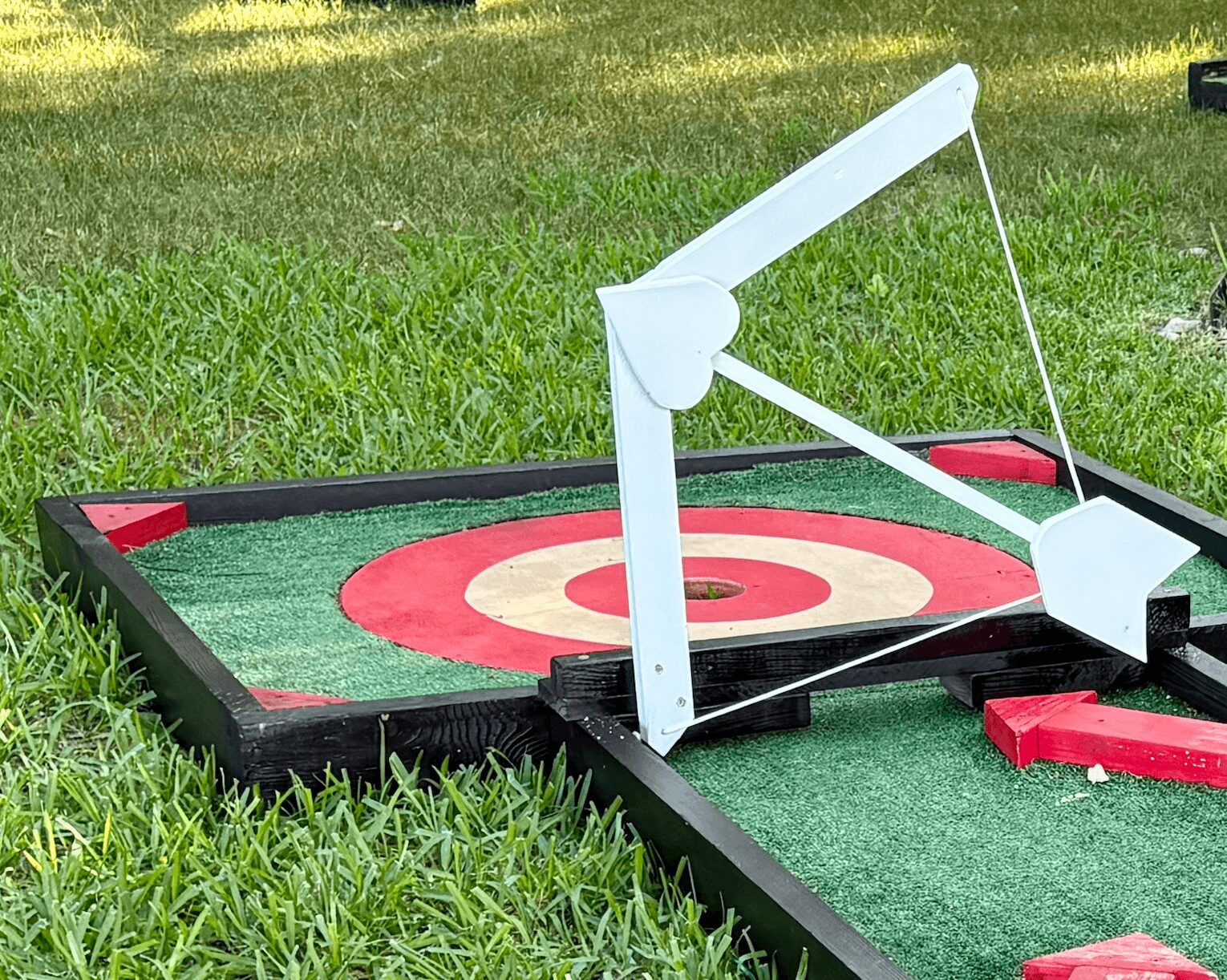 Miniature golf putting game with a white plastic putter and target on grass, with red and green accents.