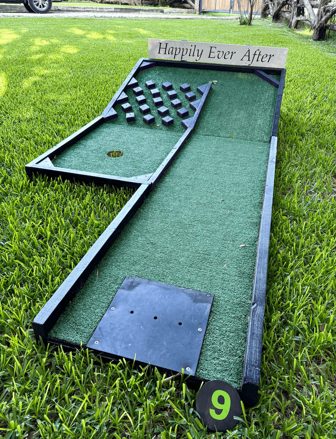 Miniature golf course with a sign reading 'Happily Ever After,' featuring a putting green, obstacles, and a hole, outdoors on grass.