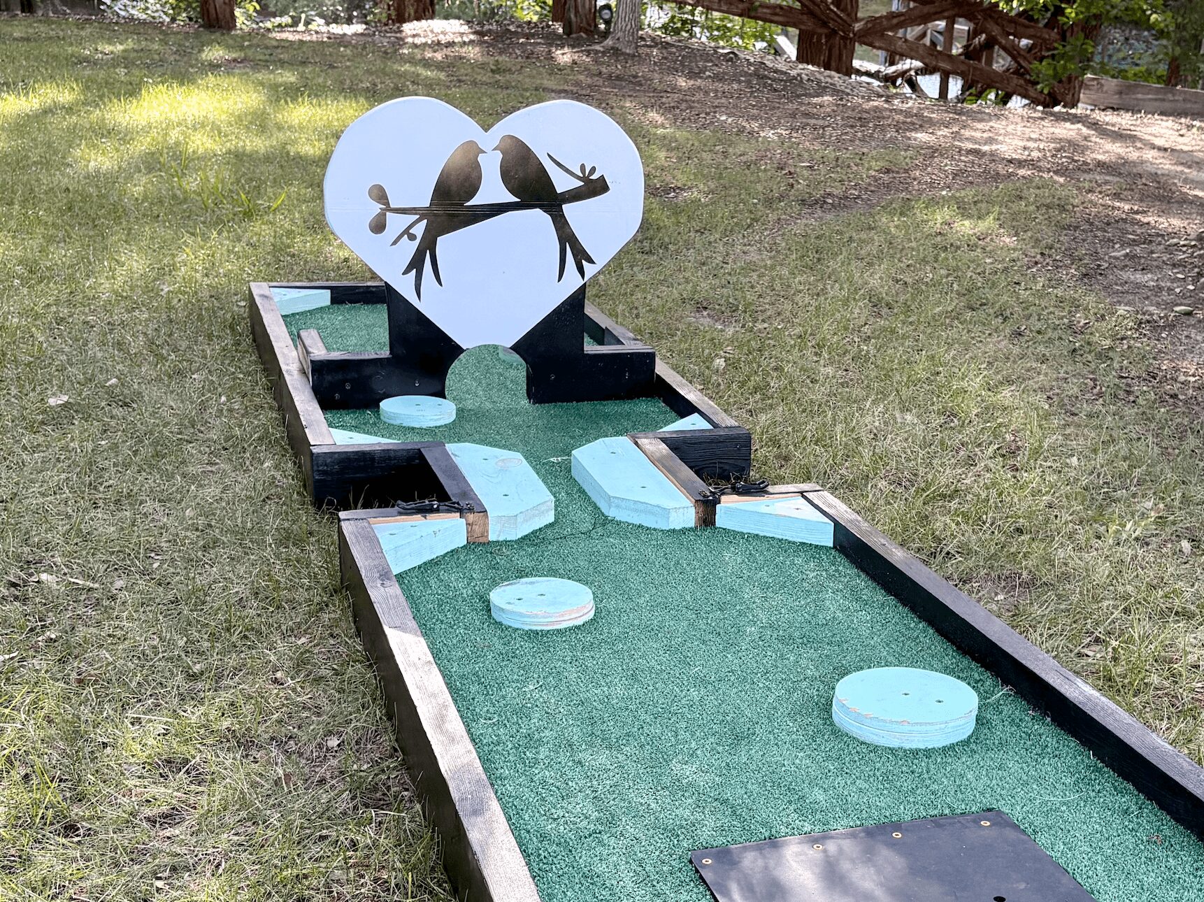 Miniature golf course with a heart-shaped decoration featuring two birds on a branch, surrounded by grass and trees.