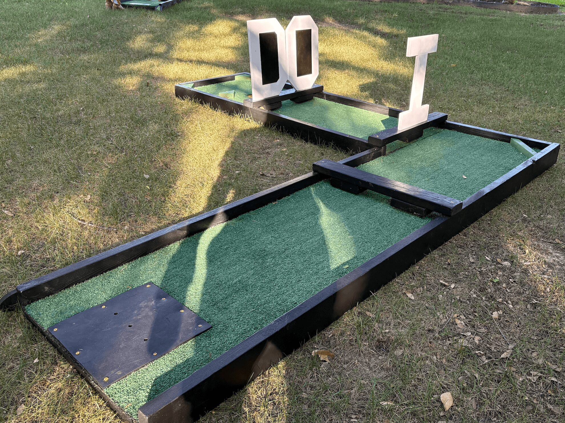Miniature golf course with two holes, one with a black square and the other with a white T-shaped obstacle, on grass.