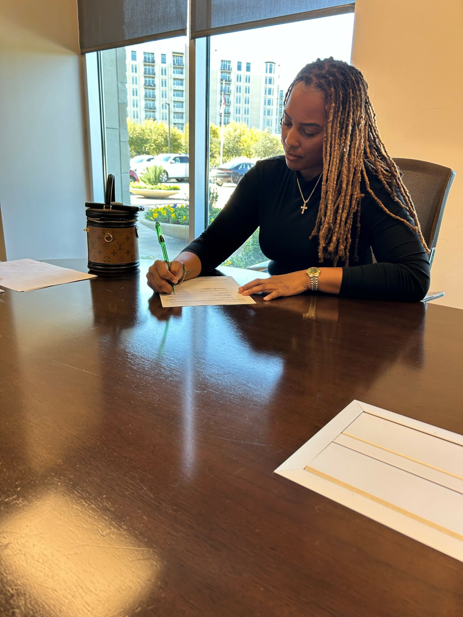 Person with long dreadlocks writing on paper at a desk in an office with large windows and city view.