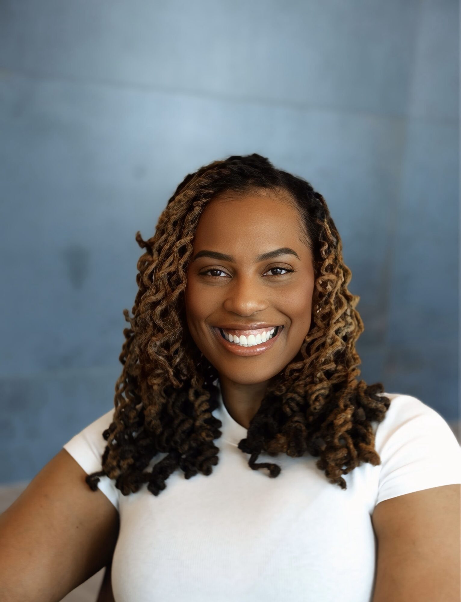 Smiling woman with curly hair wearing a white shirt against a blue background.