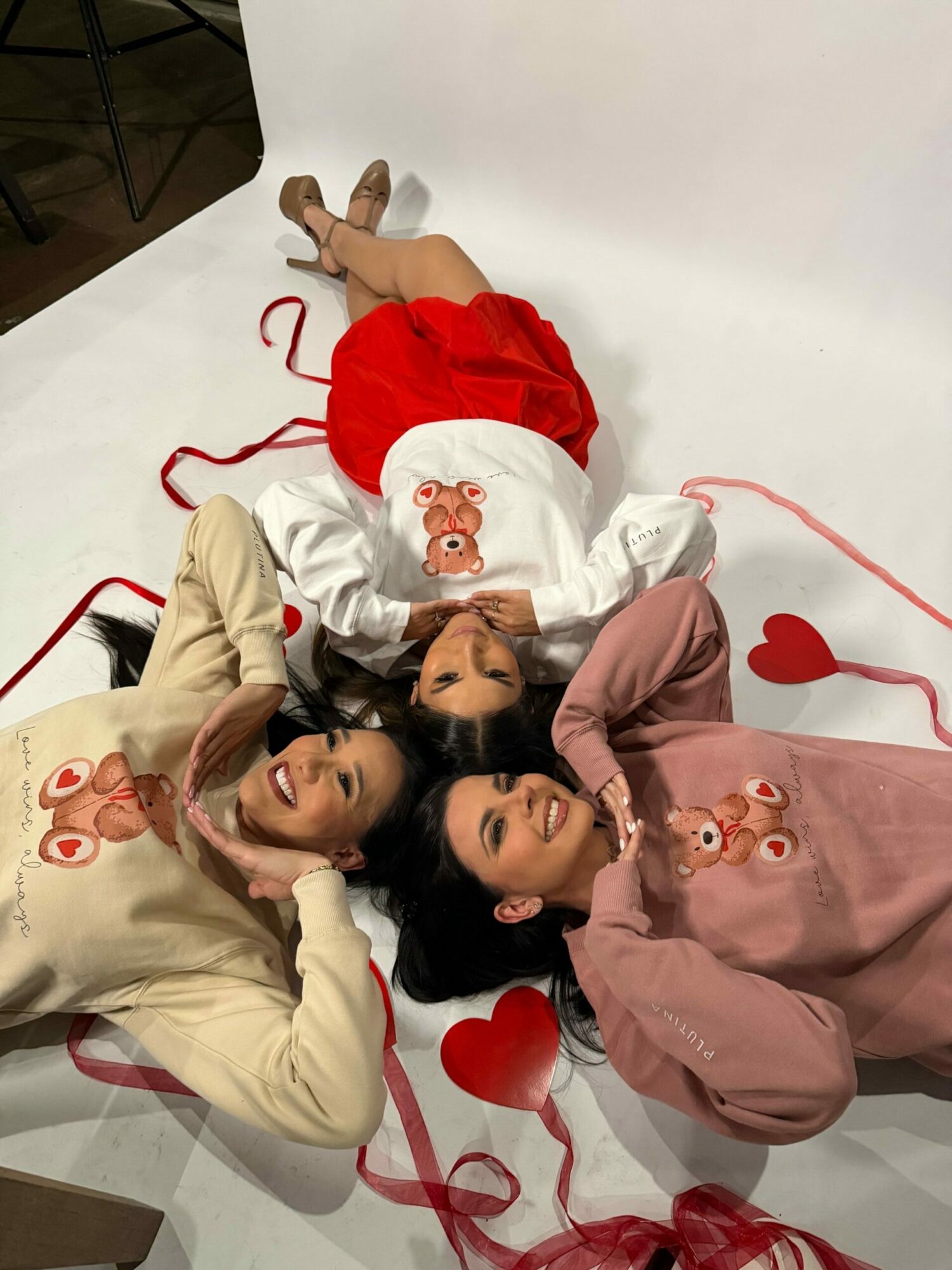 Three women lying on a white surface with red heart decorations, smiling and looking at each other.