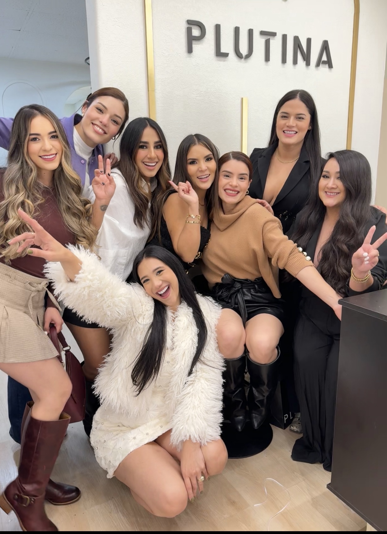 Group of eight women smiling and posing in front of a sign that says 'PLUTINA'.