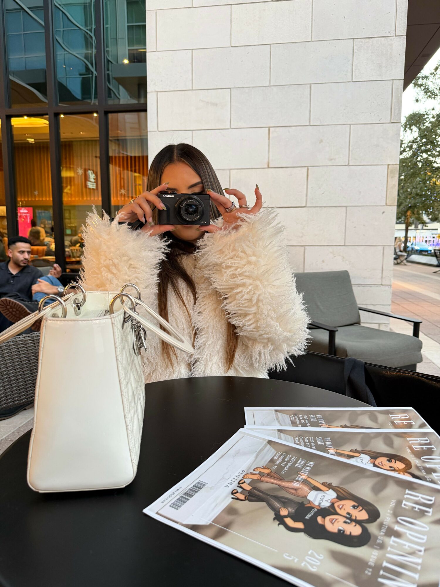 Person taking a photo with a camera, wearing a fluffy beige coat, sitting at a table with a white purse and a promotional sign, in an indoor setting.