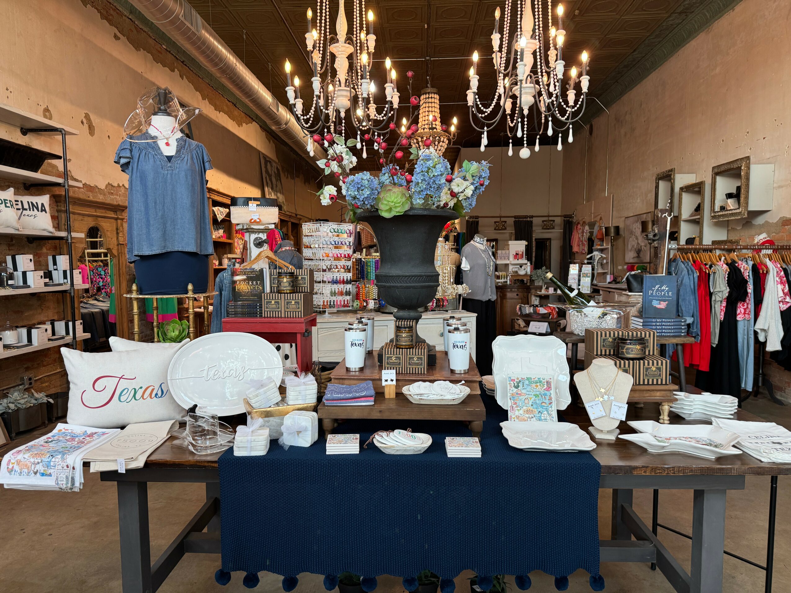 Display table with various home decor items, a large flower arrangement, and clothing racks in background.