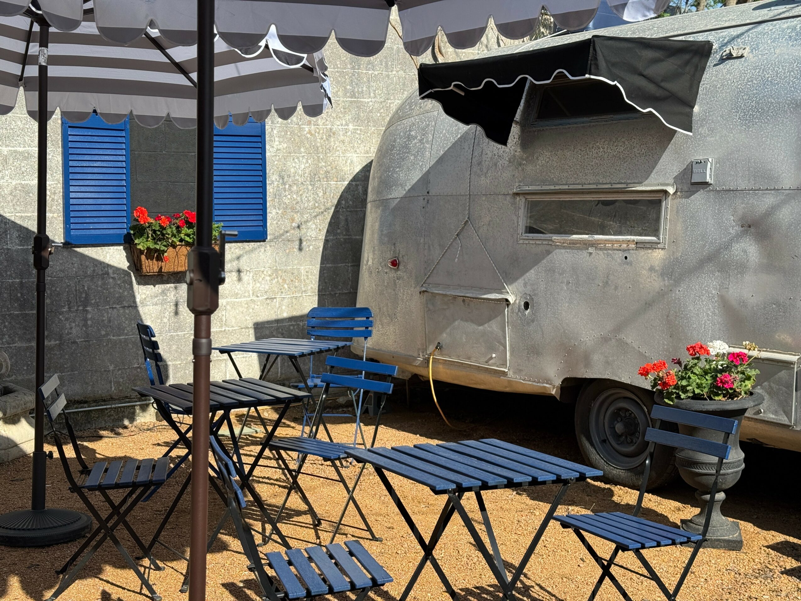 Outdoor seating area with blue chairs and tables, umbrellas, and flower pots, next to a silver trailer with a small window.