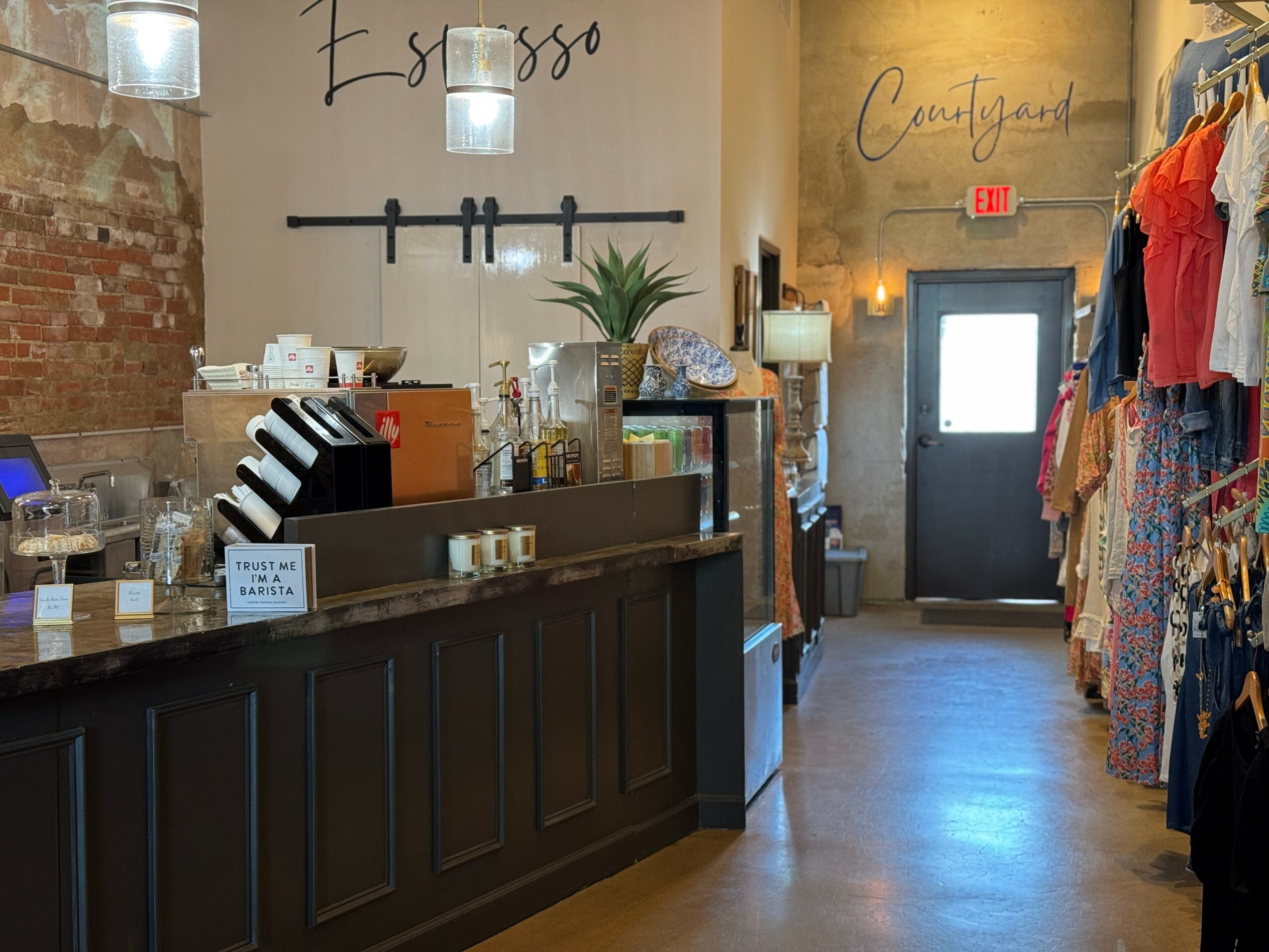 Coffee shop counter with a plant, hanging lights, and clothing racks in the background, brick wall on the left, door at the back.