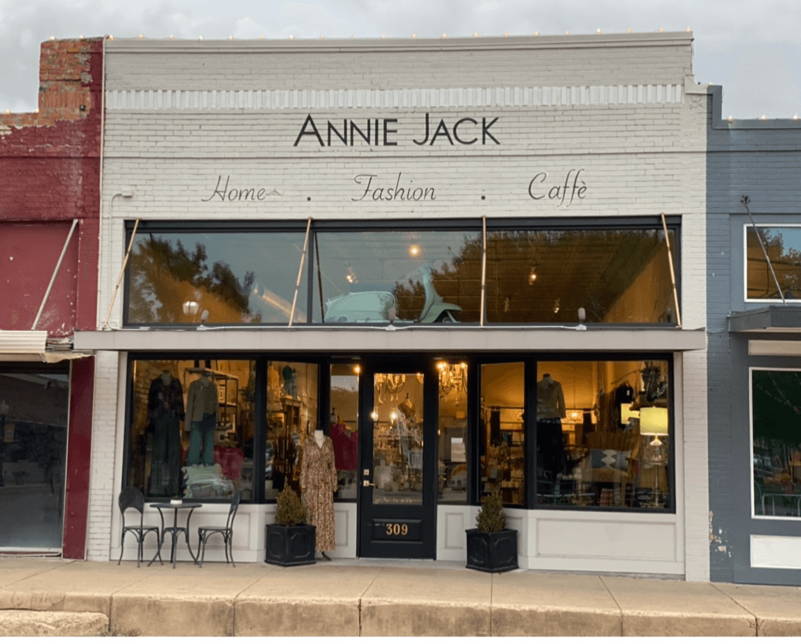 Storefront with large glass windows, displaying clothing and accessories inside, with outdoor seating and potted plants, under a white sign.