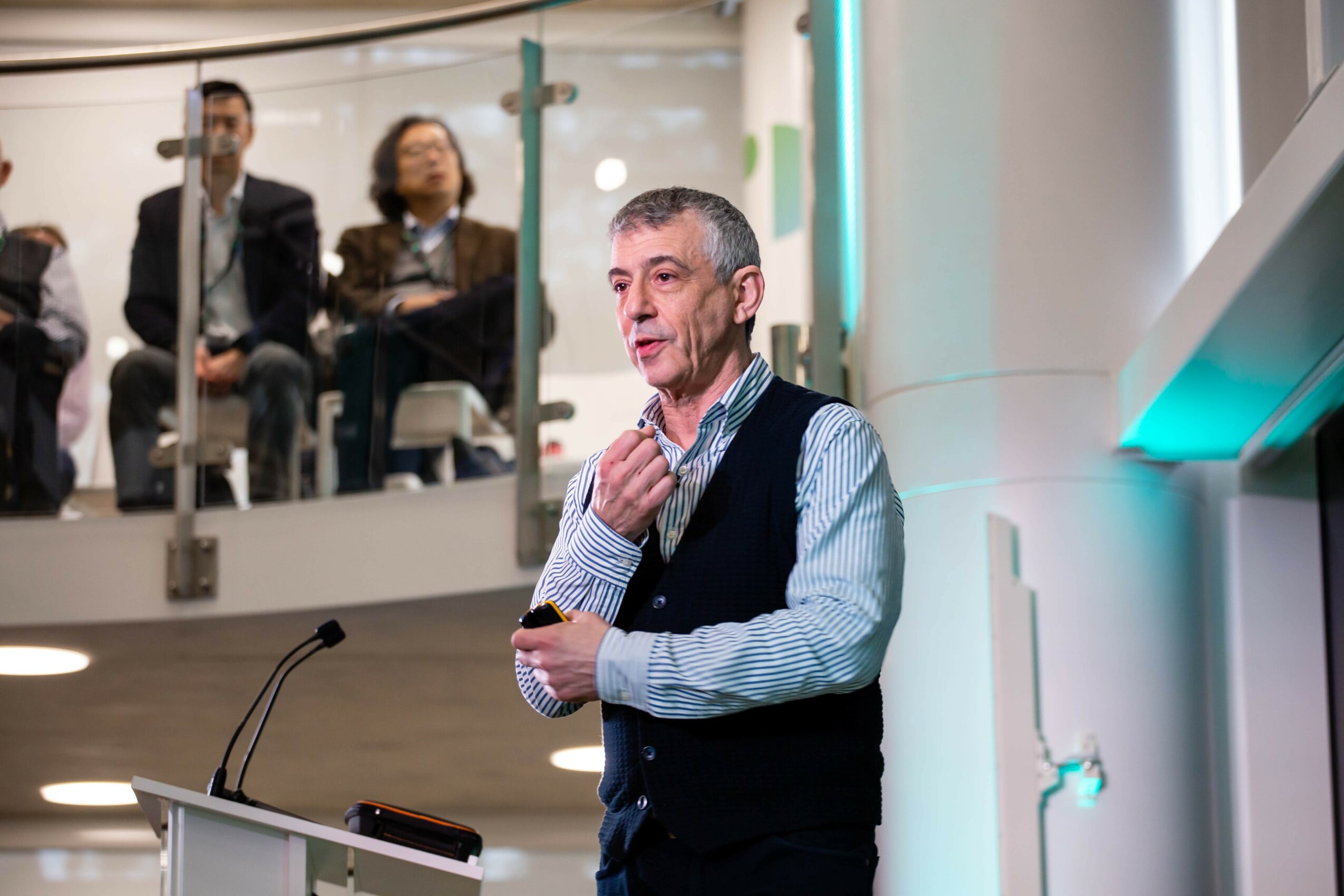 Man speaking at a conference with an audience in the background, standing near a microphone, in a modern indoor setting.