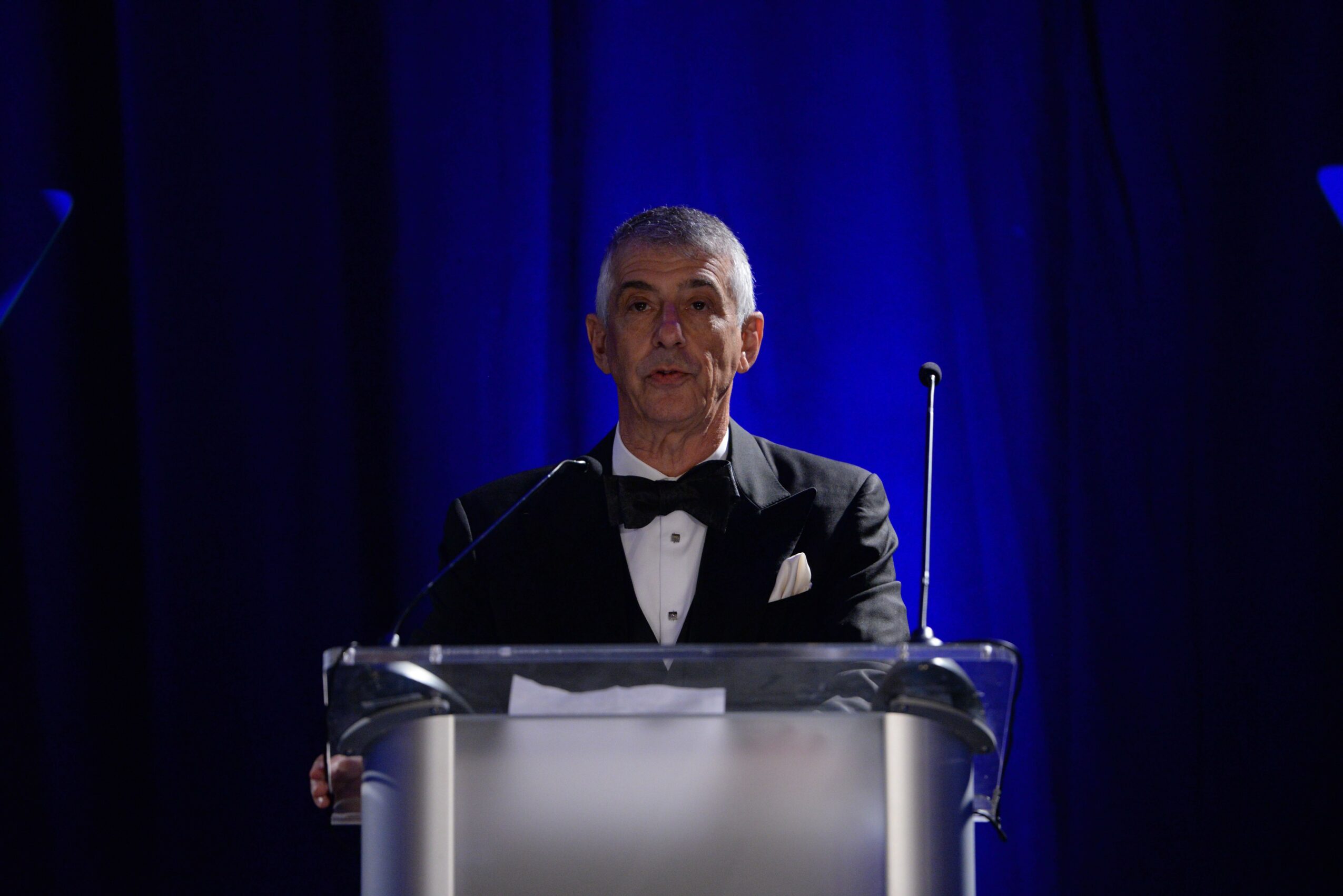 Man in tuxedo standing at a clear podium on a stage with dark blue curtains behind him.