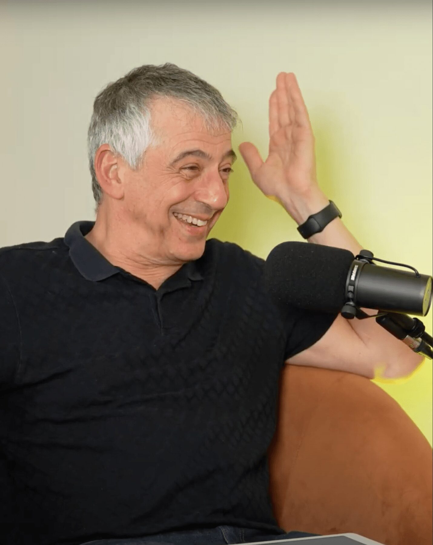 Smiling man with gray hair raising his hand, sitting in front of a microphone, against a light-colored wall.