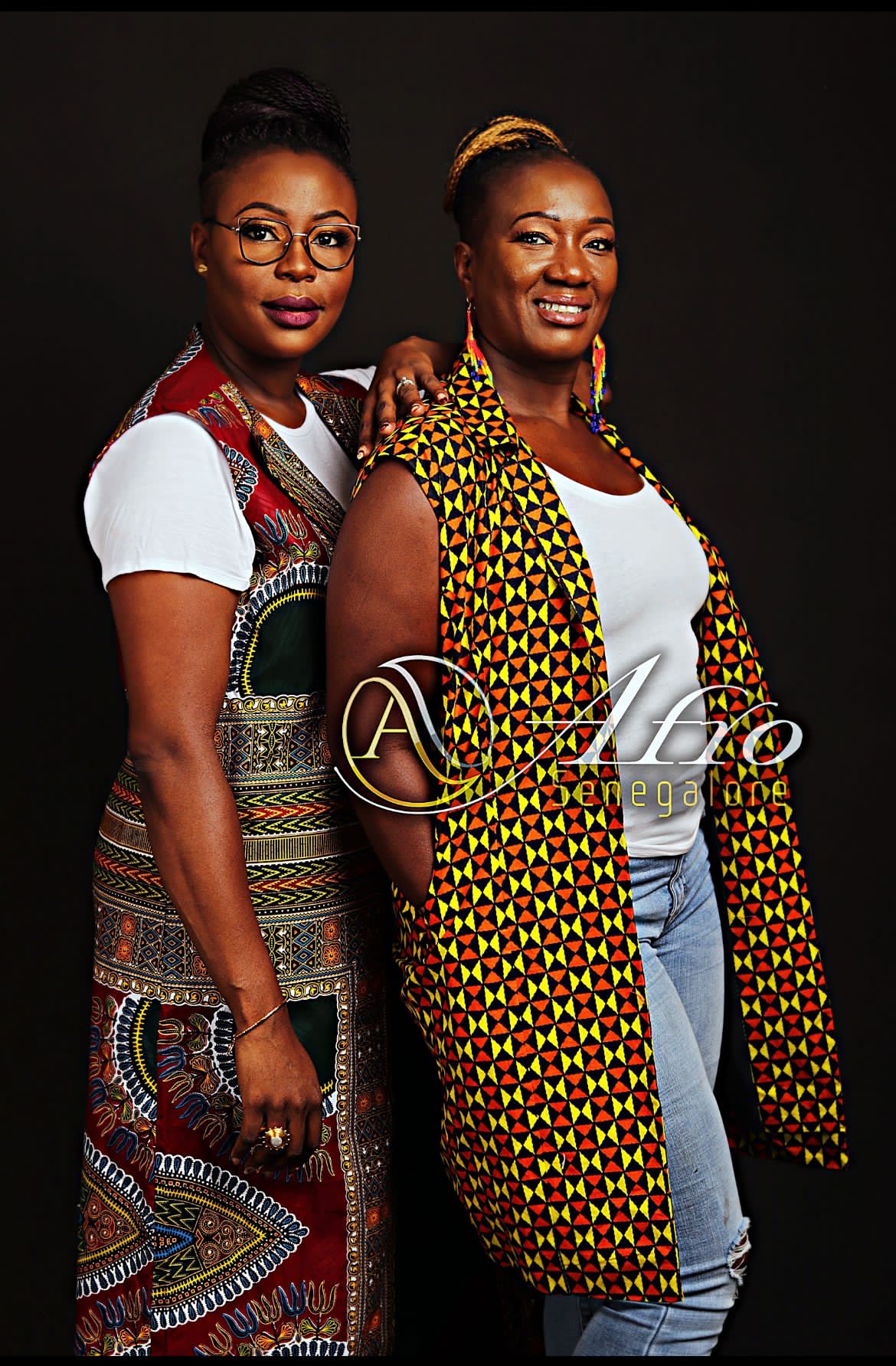 Two women stand side by side, smiling, with dark background, wearing colorful clothing and accessories.