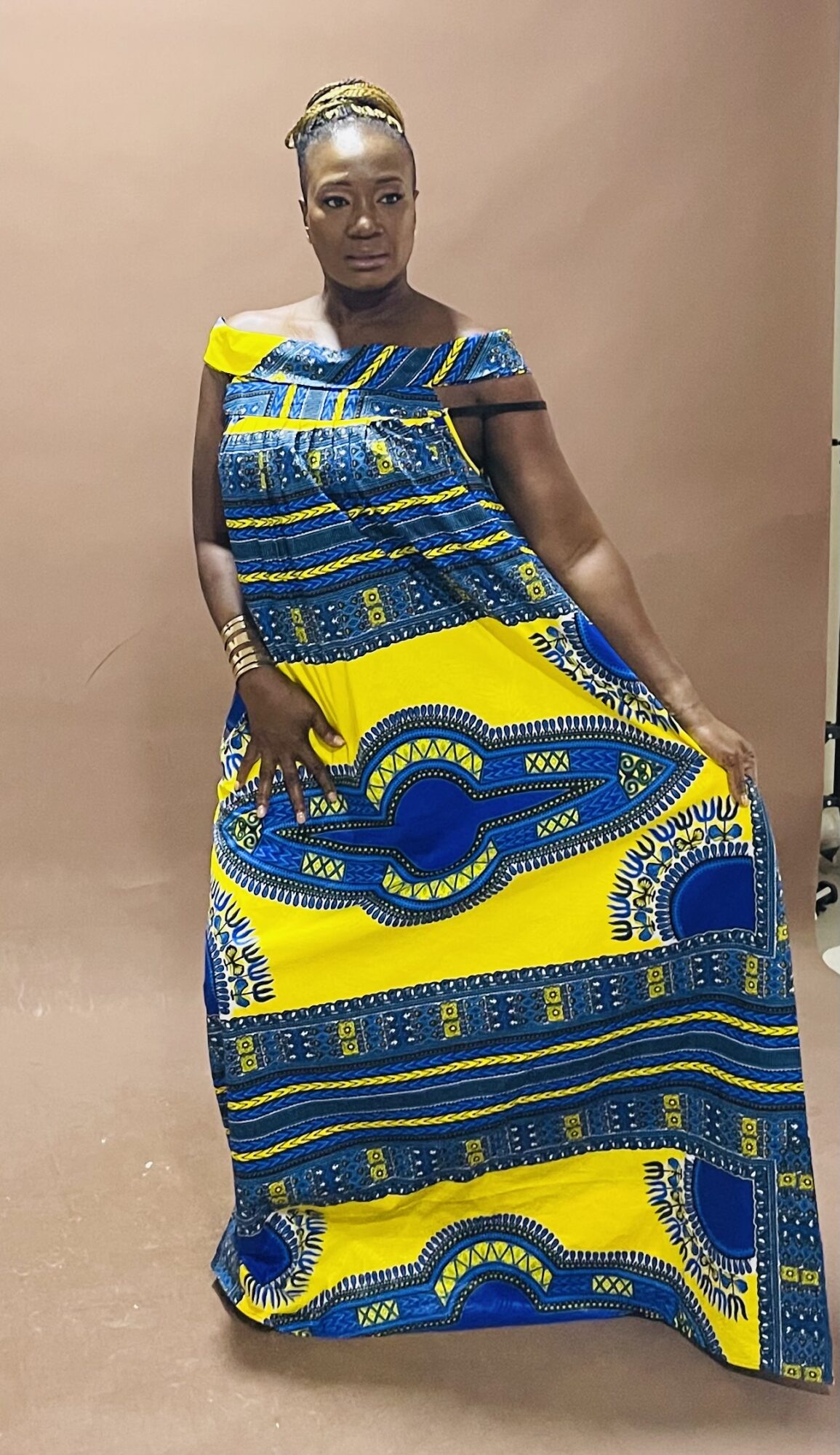 Woman in colorful patterned dress standing against a plain background, holding the dress with one hand.