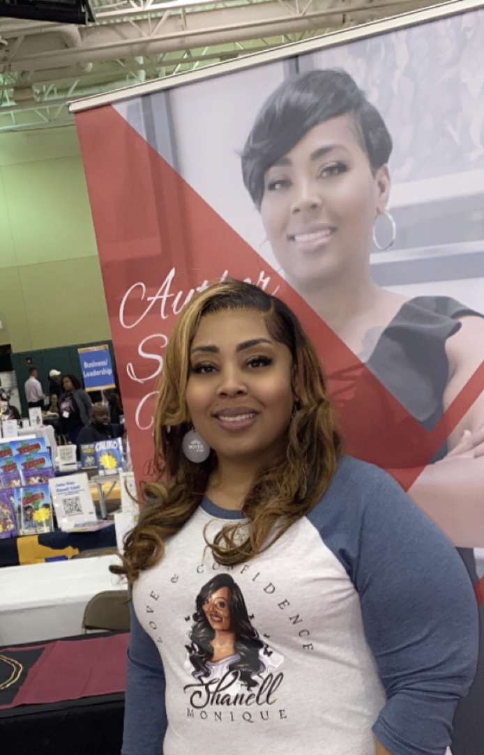 Woman with long wavy hair smiling in front of a large poster of a woman with short hair, wearing a white and blue shirt.