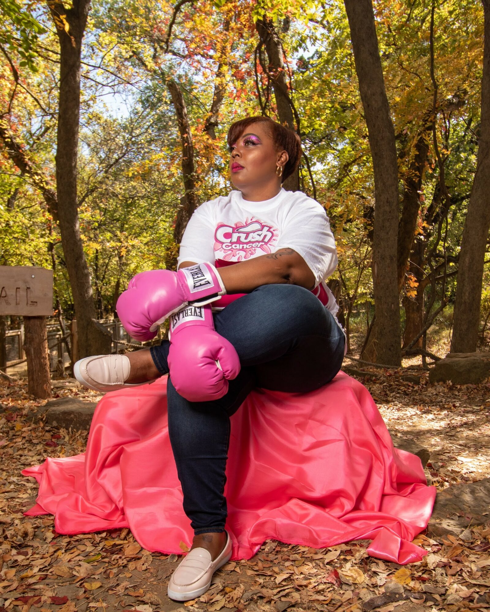 Woman sitting on a pink cloth in a forest with autumn leaves, wearing boxing gloves and casual clothes.