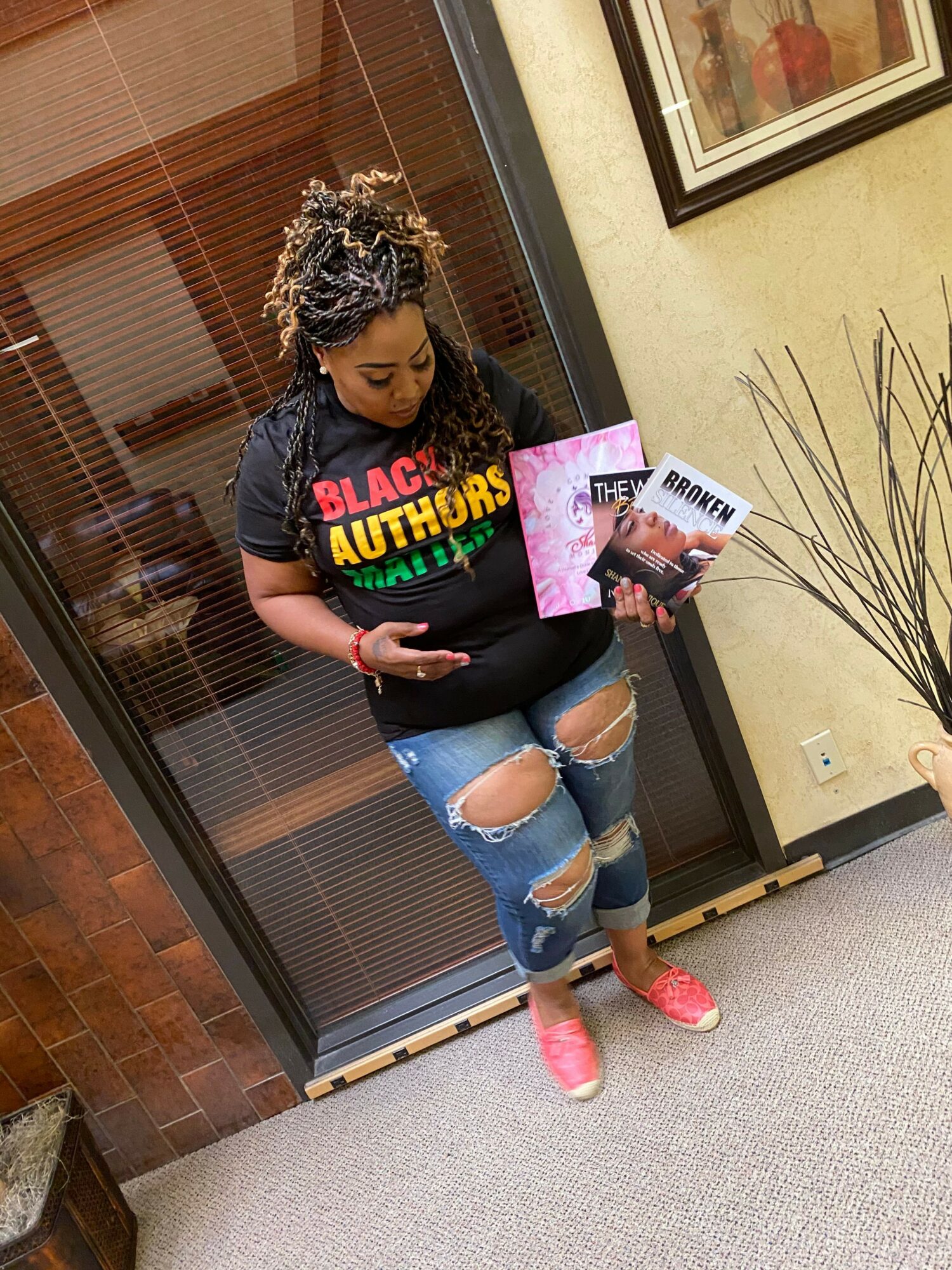 Person standing in front of a glass door, holding a magazine, wearing ripped jeans and pink shoes.