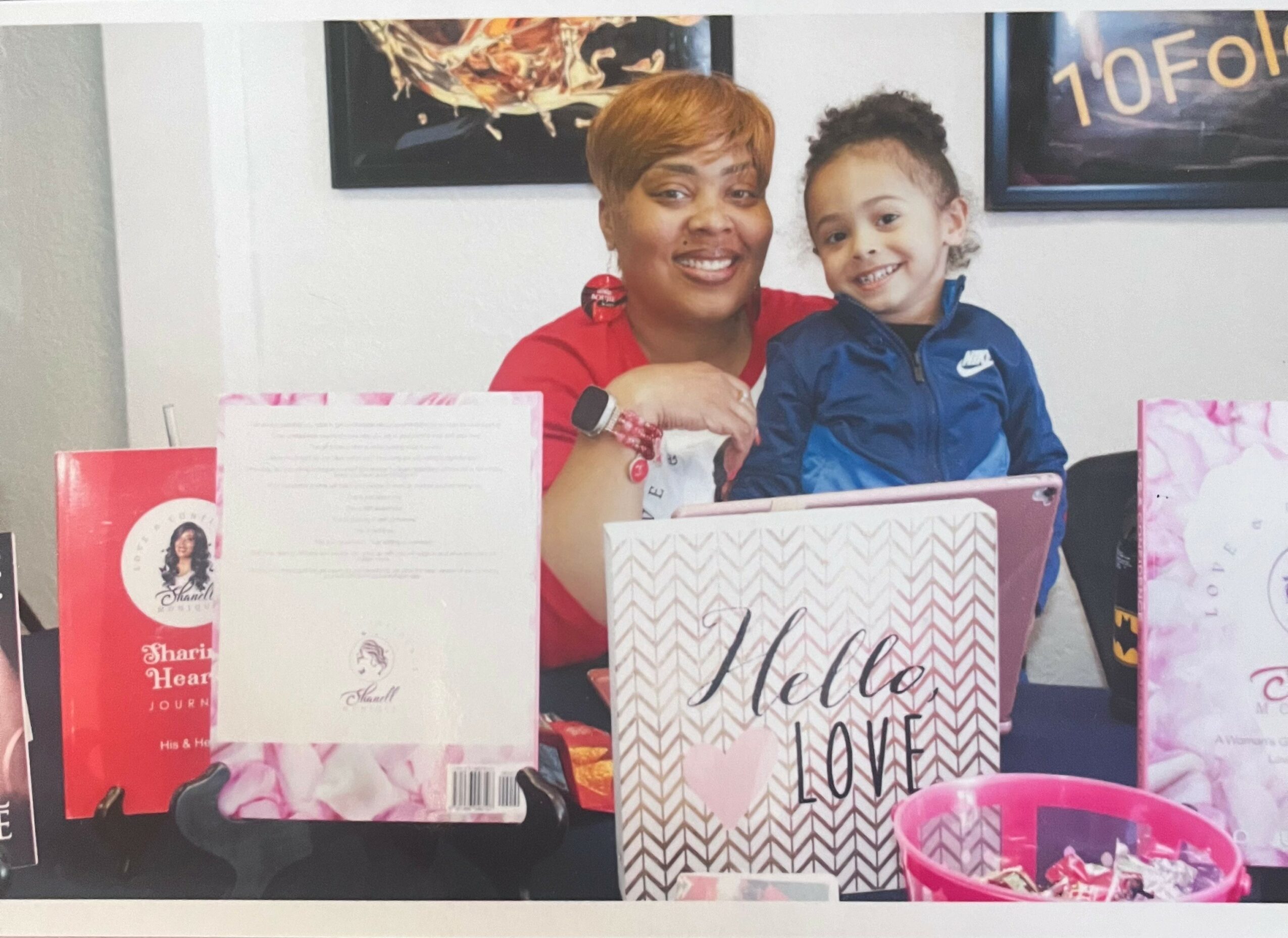 Woman and girl smiling behind a table with greeting cards and pink items, indoors.