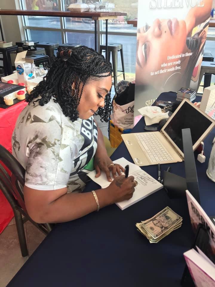 Woman writing in a notebook at a table with a laptop, cash, and promotional poster in background.