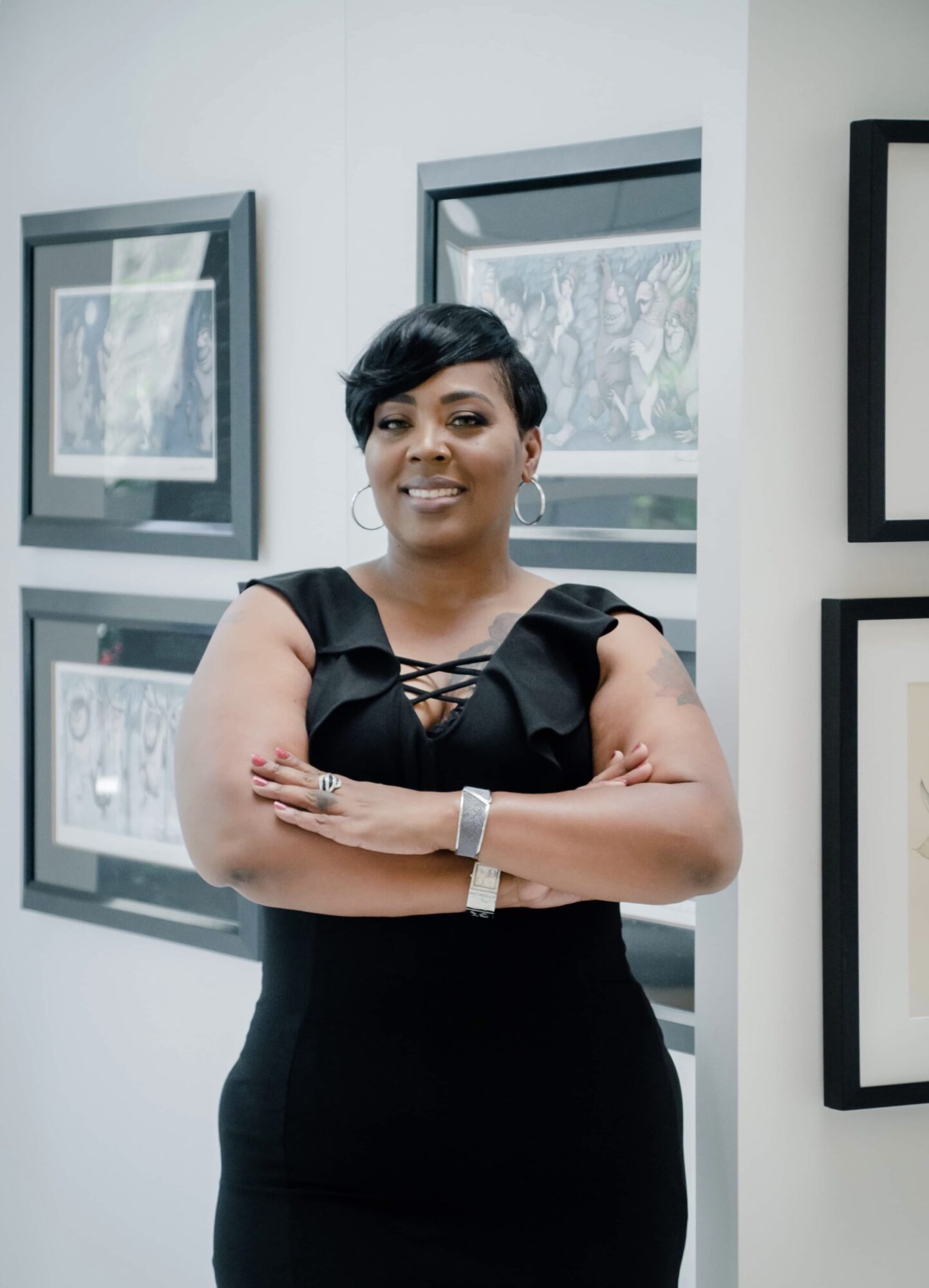Woman with short black hair wearing a black dress with ruffled sleeves, standing with arms crossed in an art gallery.