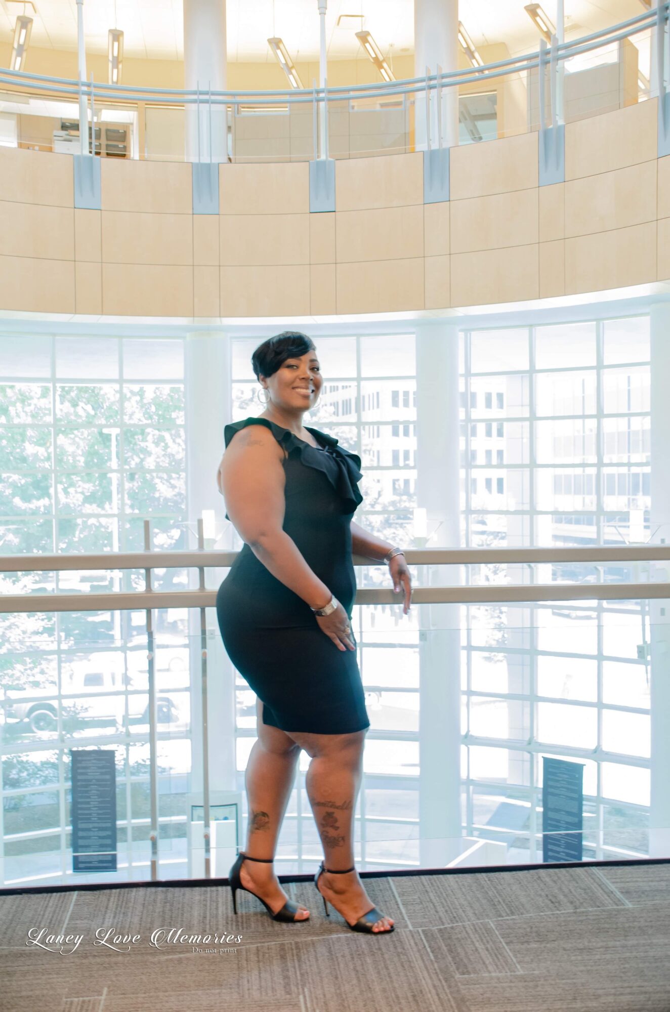 Woman in black dress and heels standing by a railing in a bright, modern building with large windows.