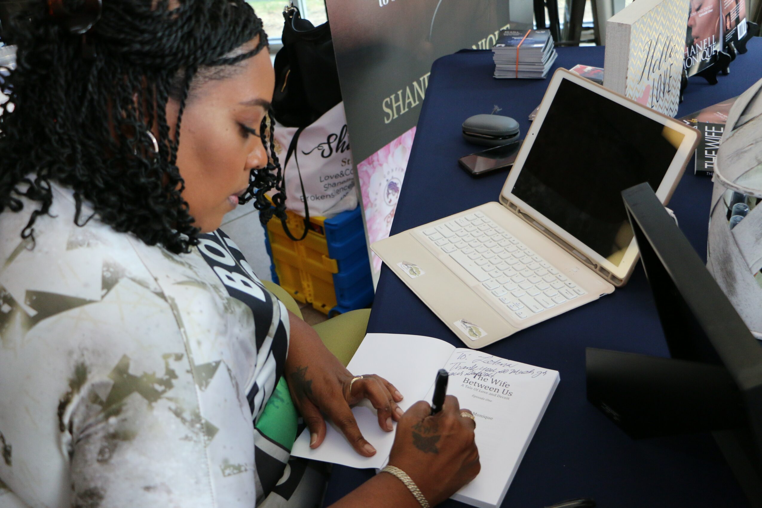 Woman with curly hair writing in a notebook at a table with a laptop and electronic devices.