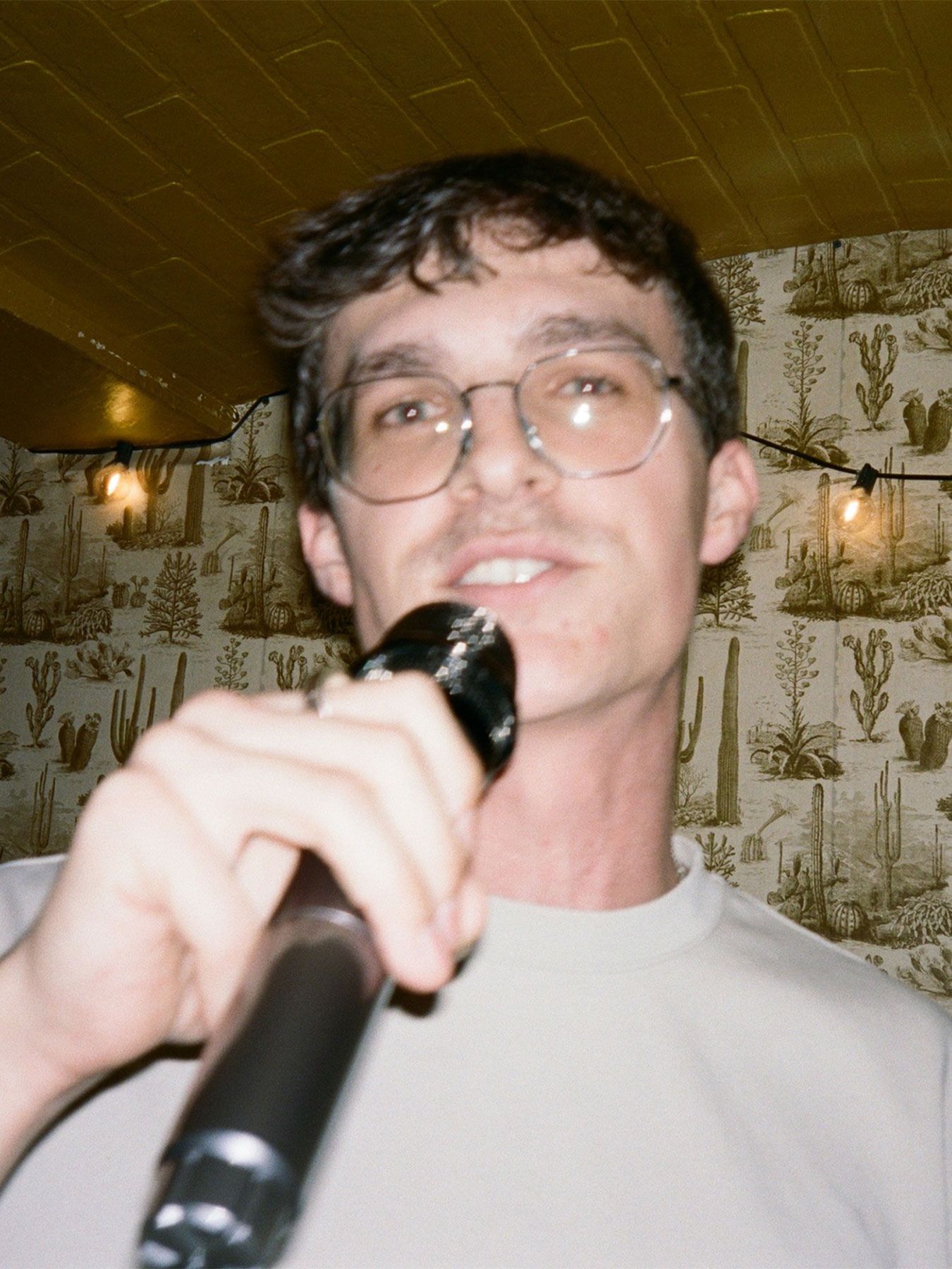 Young man with glasses holding a microphone, smiling, in a decorated room with patterned wallpaper and warm lighting.