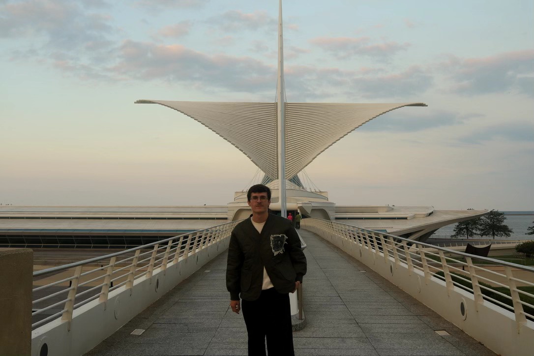 Person standing on a bridge with a modern architectural structure in the background, sky with clouds, and water nearby.