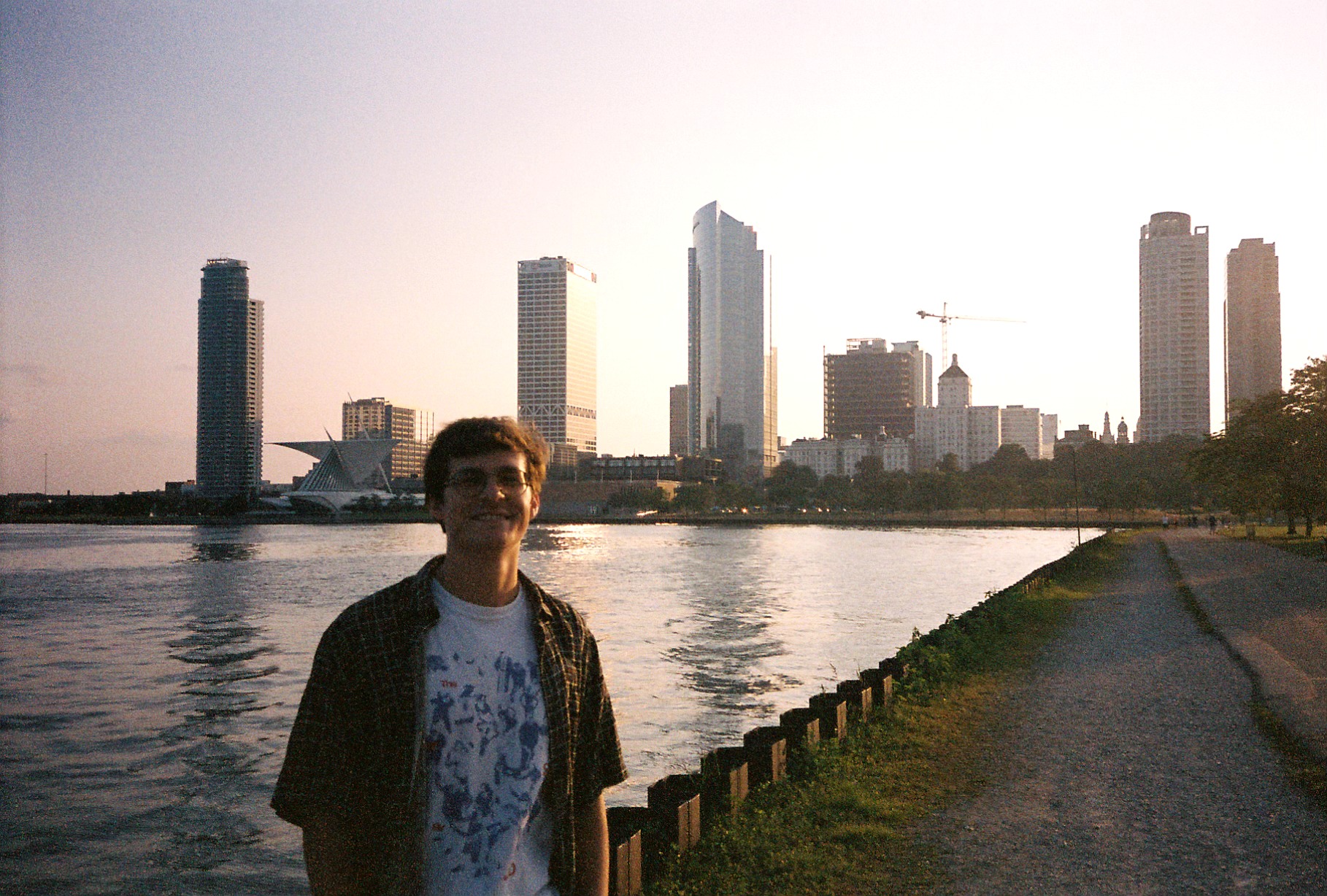 Person standing near water with city skyline and tall buildings in the background during sunset.