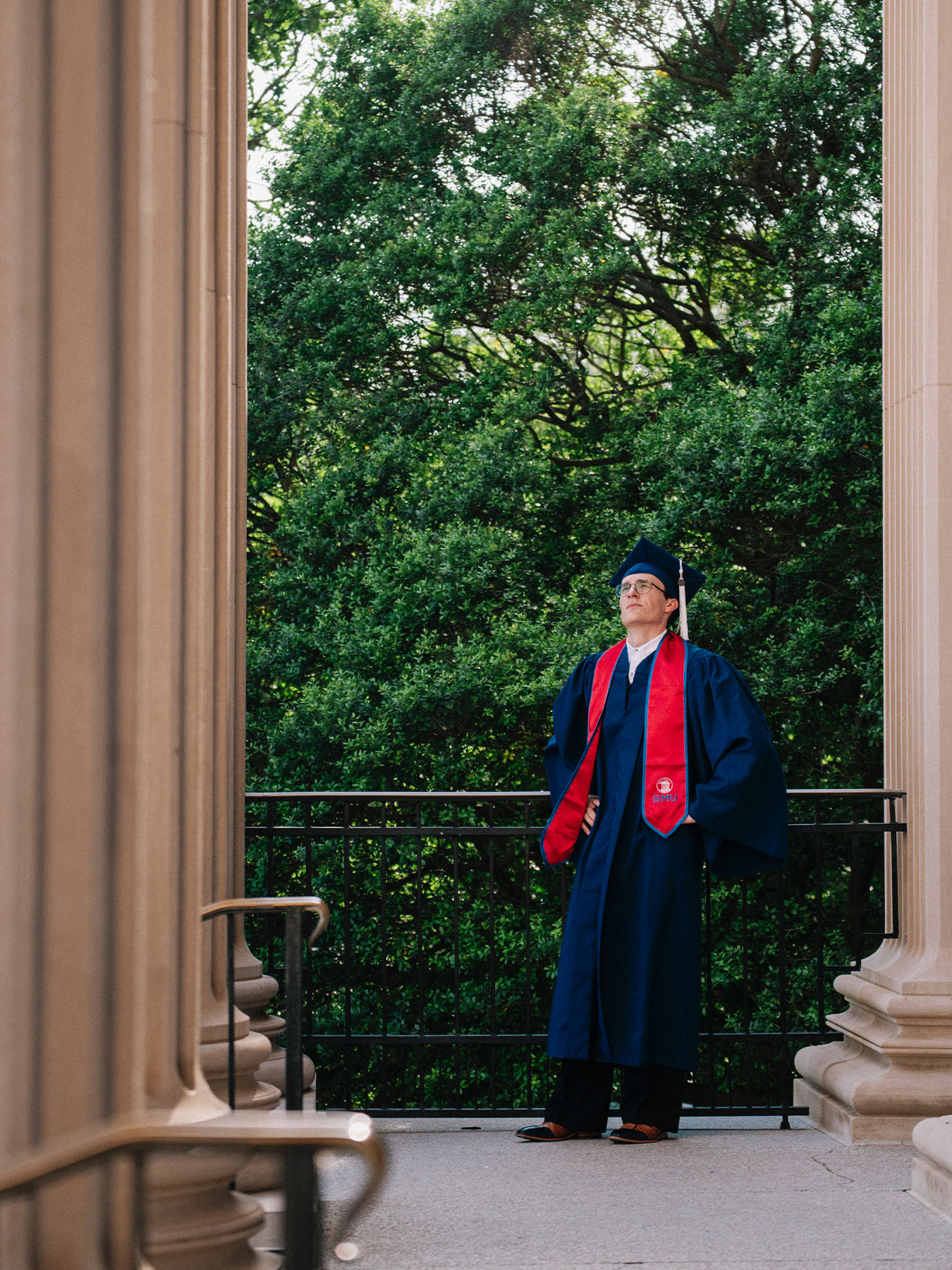 Person in graduation gown and cap standing on balcony with green trees behind, framed by columns.