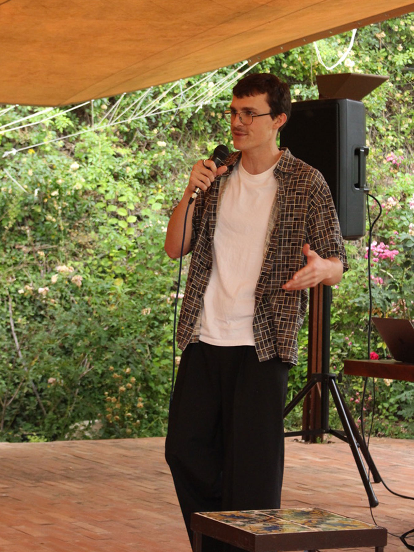 Young man speaking into a microphone outdoors, with trees and greenery in the background.