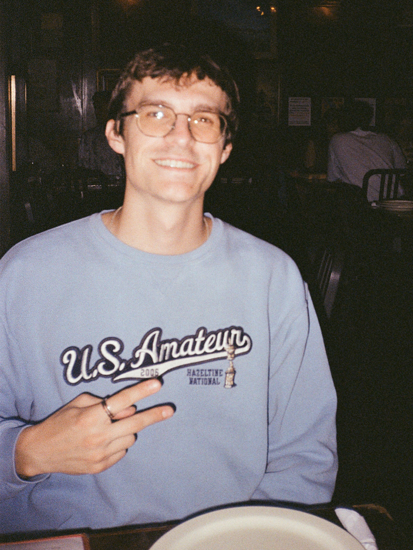 Young man with glasses smiling, wearing a light blue sweatshirt with text, sitting at a table in a dimly lit setting.