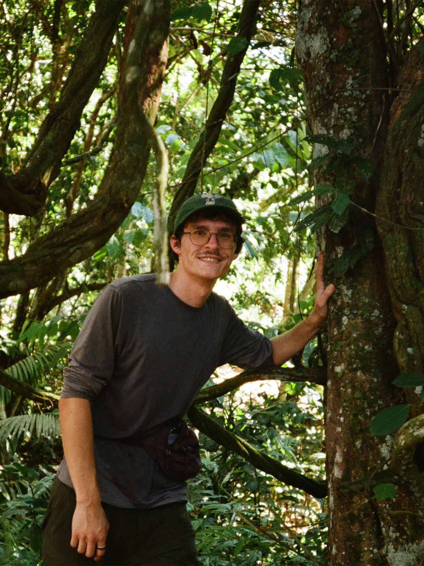 Person with glasses and a cap smiling, standing in a dense forest, touching a tree trunk.