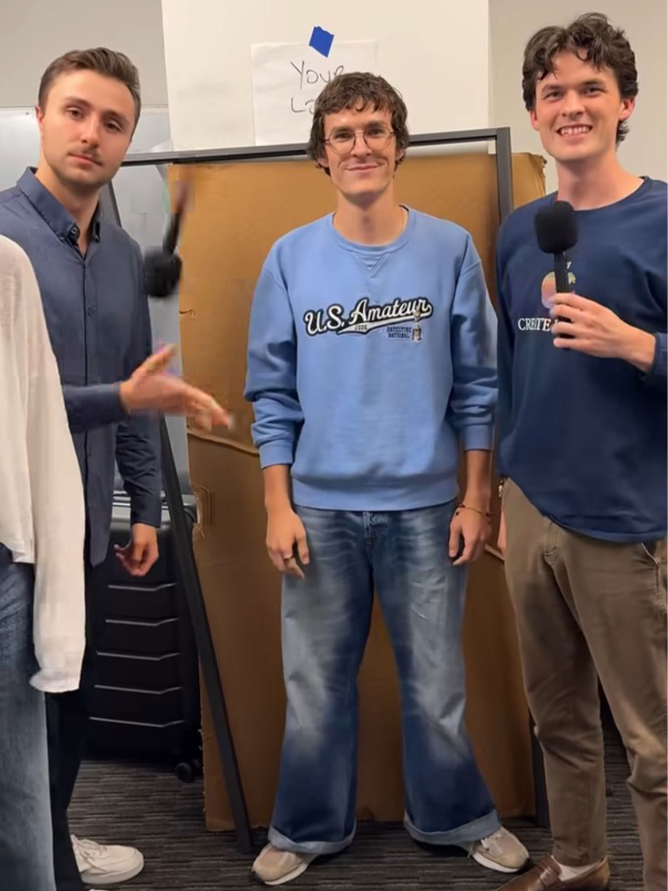 Three young men standing indoors, two holding microphones, smiling, with a whiteboard behind them.