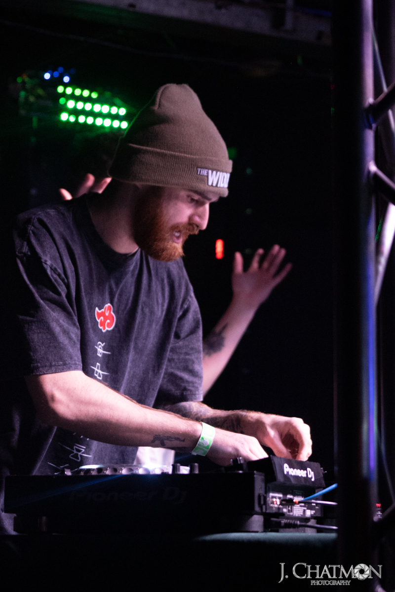 Person wearing a beanie and dark shirt, operating DJ equipment with a hand raised in the background, colorful lights behind.