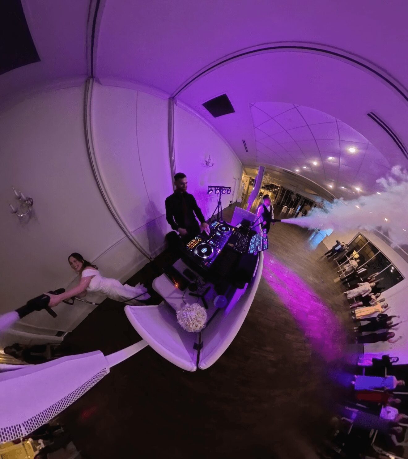 DJ playing music at an event with a child lying on a table, purple lighting, and smoke effects, curved ceiling, and guests in background.