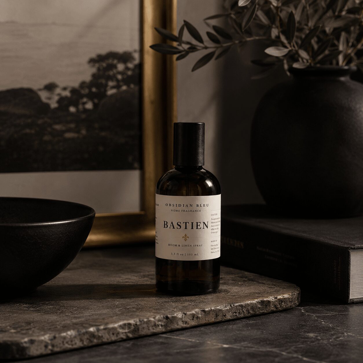 A dark bottle labeled 'BASTIEN' on a textured surface with a black bowl, framed picture, and plant in the background.